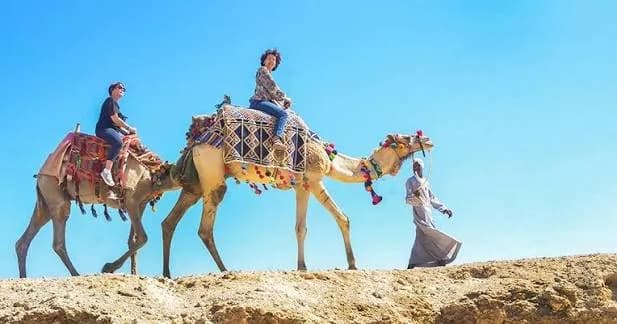 Two guests riding camels along a rocky desert ridge with an Egyptian handler walking beside them