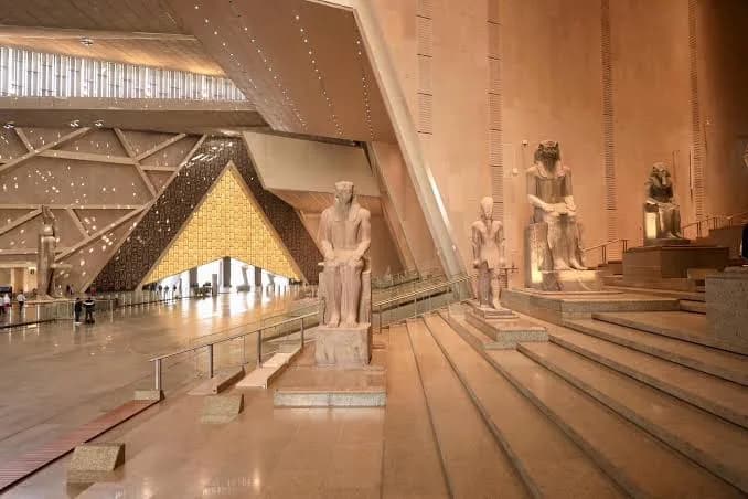 Grand Egyptian Museum staircase with seated pharaoh statues and the museum's pyramid-shaped window behind